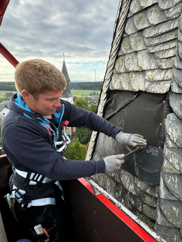 Dachdecker von Meyendriesch-Dach bei der Reparatur einer Schieferfassade an einem Kirchturm – gesicherter Einsatz in der Höhe mit Blick auf eine Hunsrücker Ortschaft