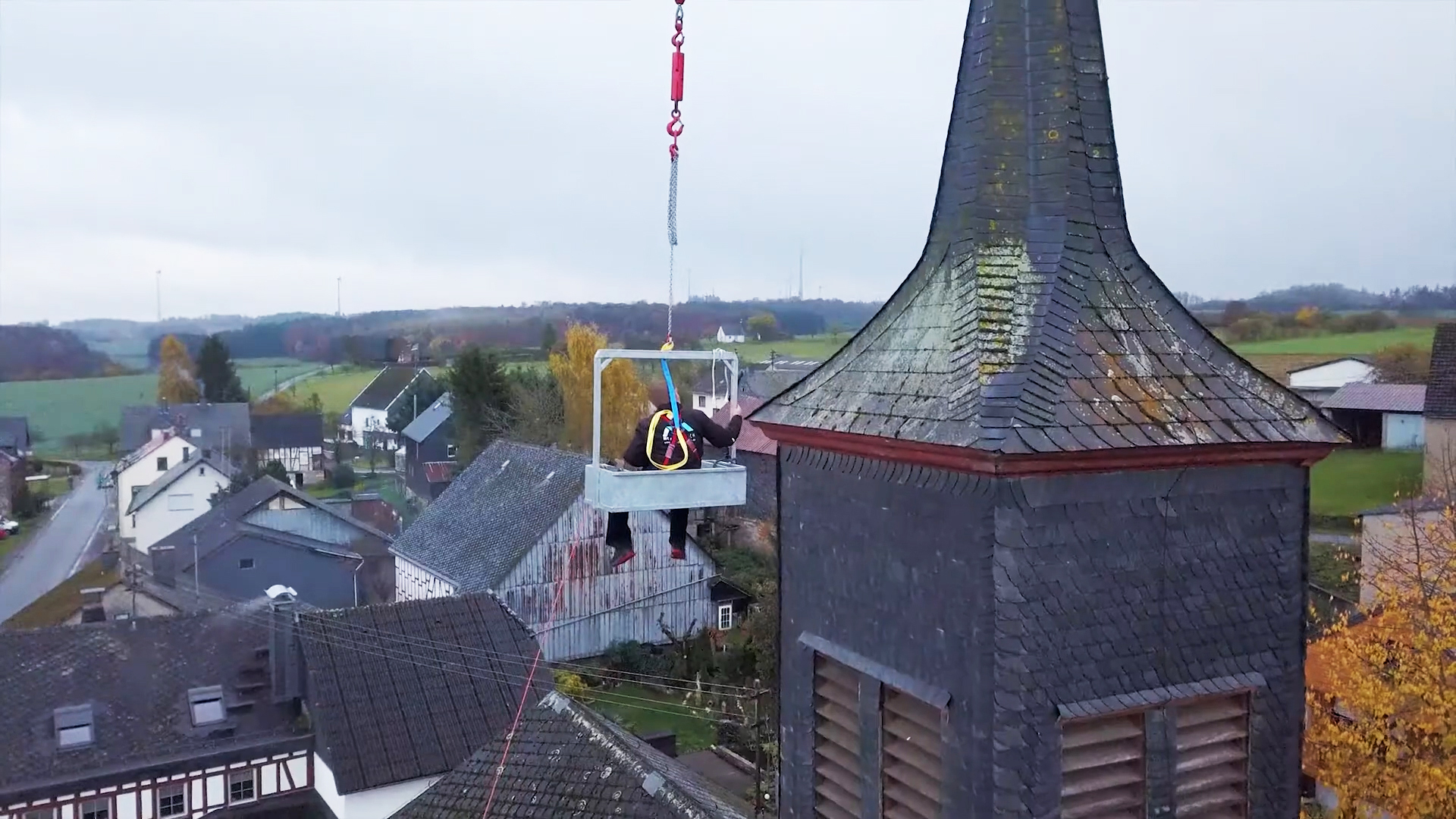 Mitarbeiter von Meyendriesch-Dach bei Wartungs- und Reparaturarbeiten am Schieferdach eines Kirchturms – Einsatz per Krankorb über einem Hunsrücker Dorf im Herbst