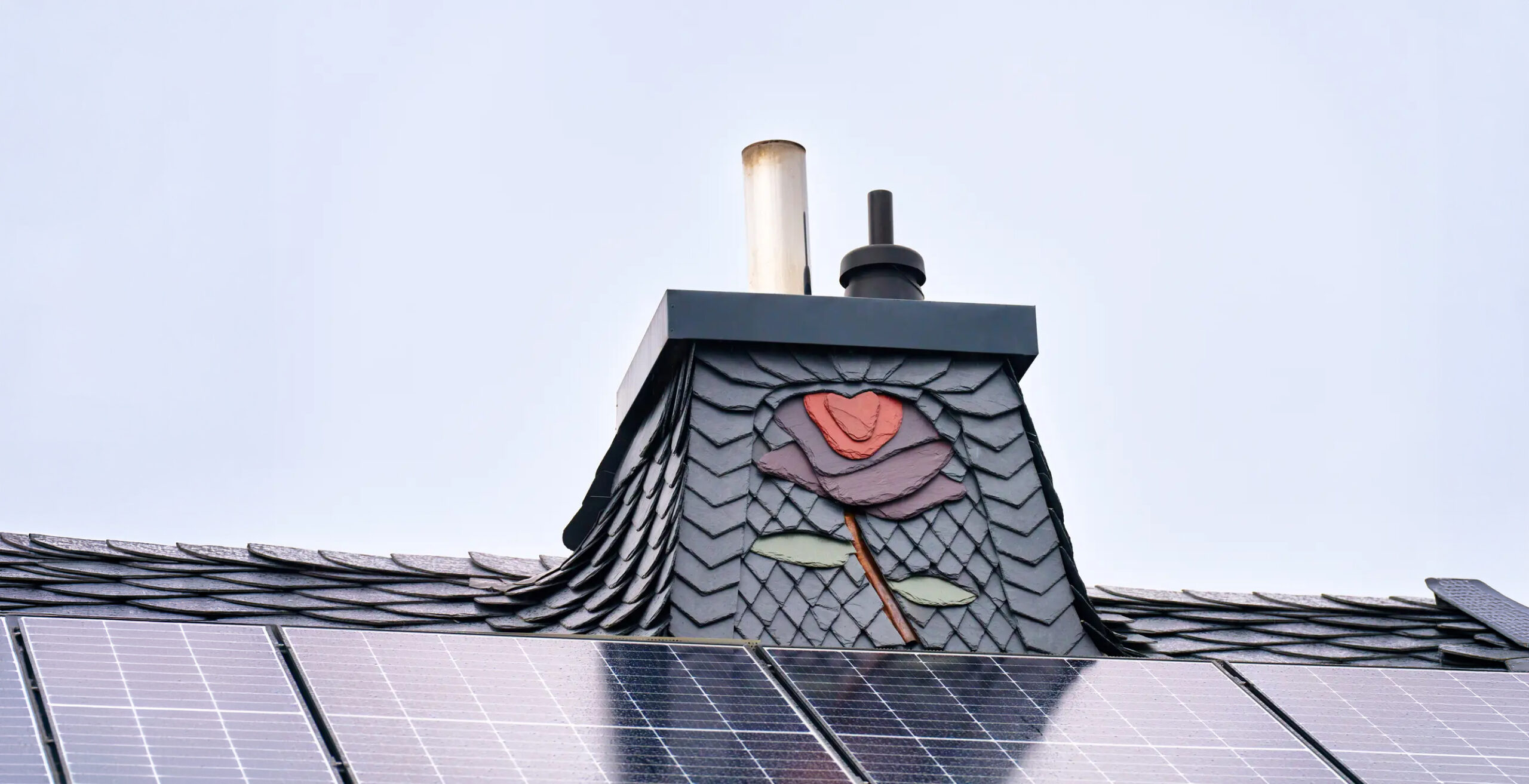In Schiefer verkleideter Schornstein mit farbigem Rosen-Ornament auf einem Schieferdach mit Photovoltaikanlage – Schieferkunst von Meyendriesch-Dach, Hunsrück
