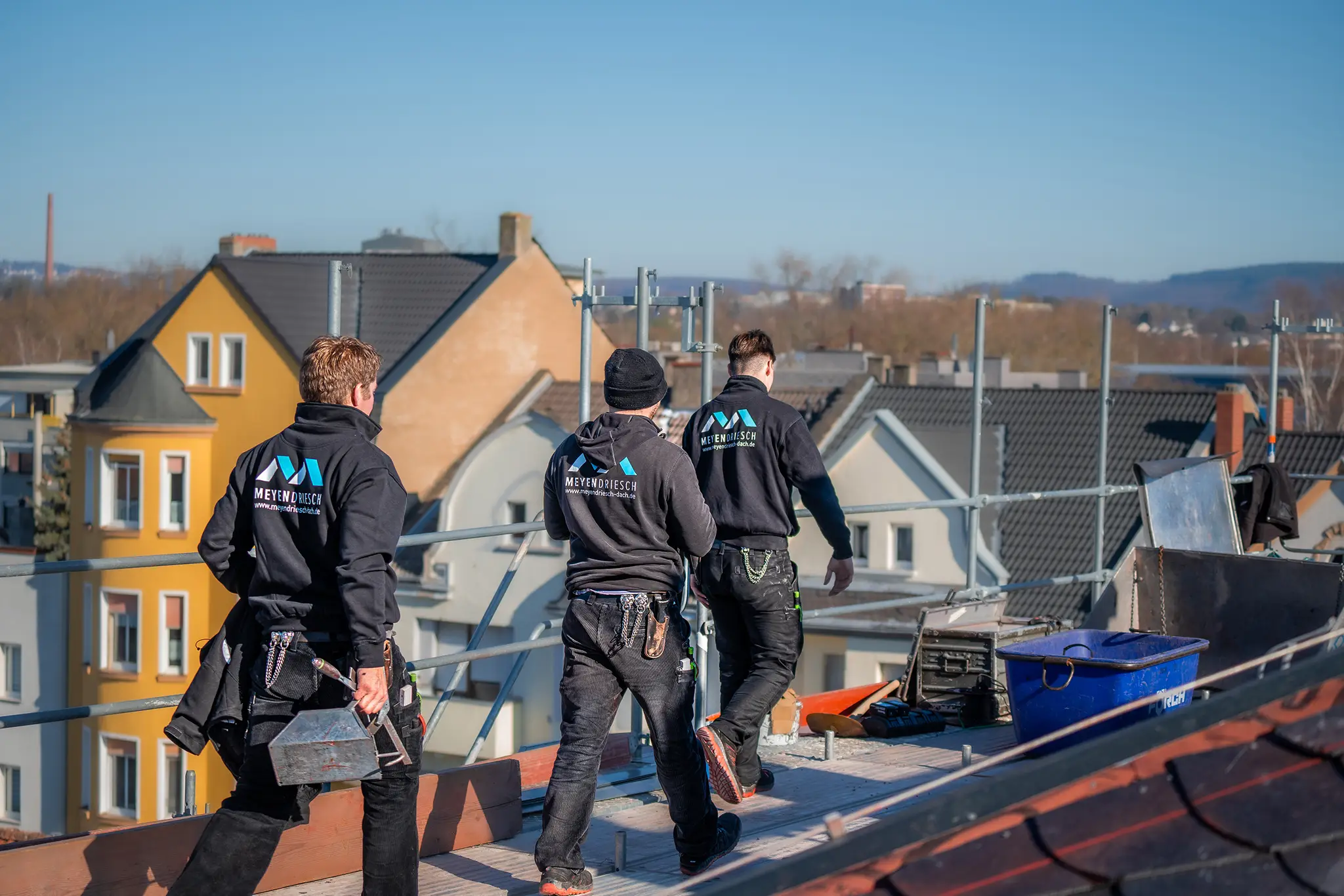 Drei Mitarbeiter von Meyendriesch-Dach in Arbeitskleidung auf einem Flachdach mit Blick über die Dächer einer Stadt – das Team von Meyendriesch-Dach, Hunsrück