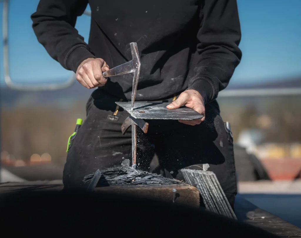 Dachdecker von Meyendriesch-Dach beim handwerklichen Zuschneiden einer Schieferplatte mit Schieferhammer und Amboss – traditionelles Schieferhandwerk auf der Baustelle