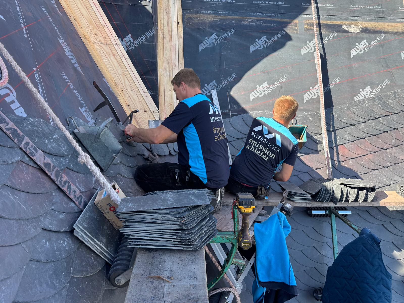 Zwei Mitarbeiter von Meyendriesch-Dach beim gemeinsamen Verlegen von Schieferplatten im Bogenschnitt auf einer Baustelle – #missionschiefer, Hunsrück